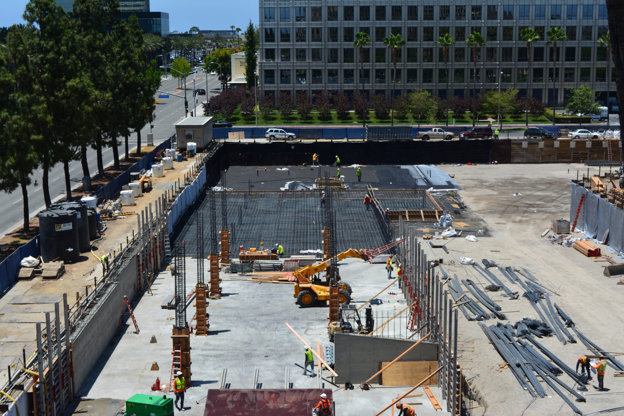 This job site features various metal bars. There’s a road to the left of the image with tall, green trees along the sidewalk.