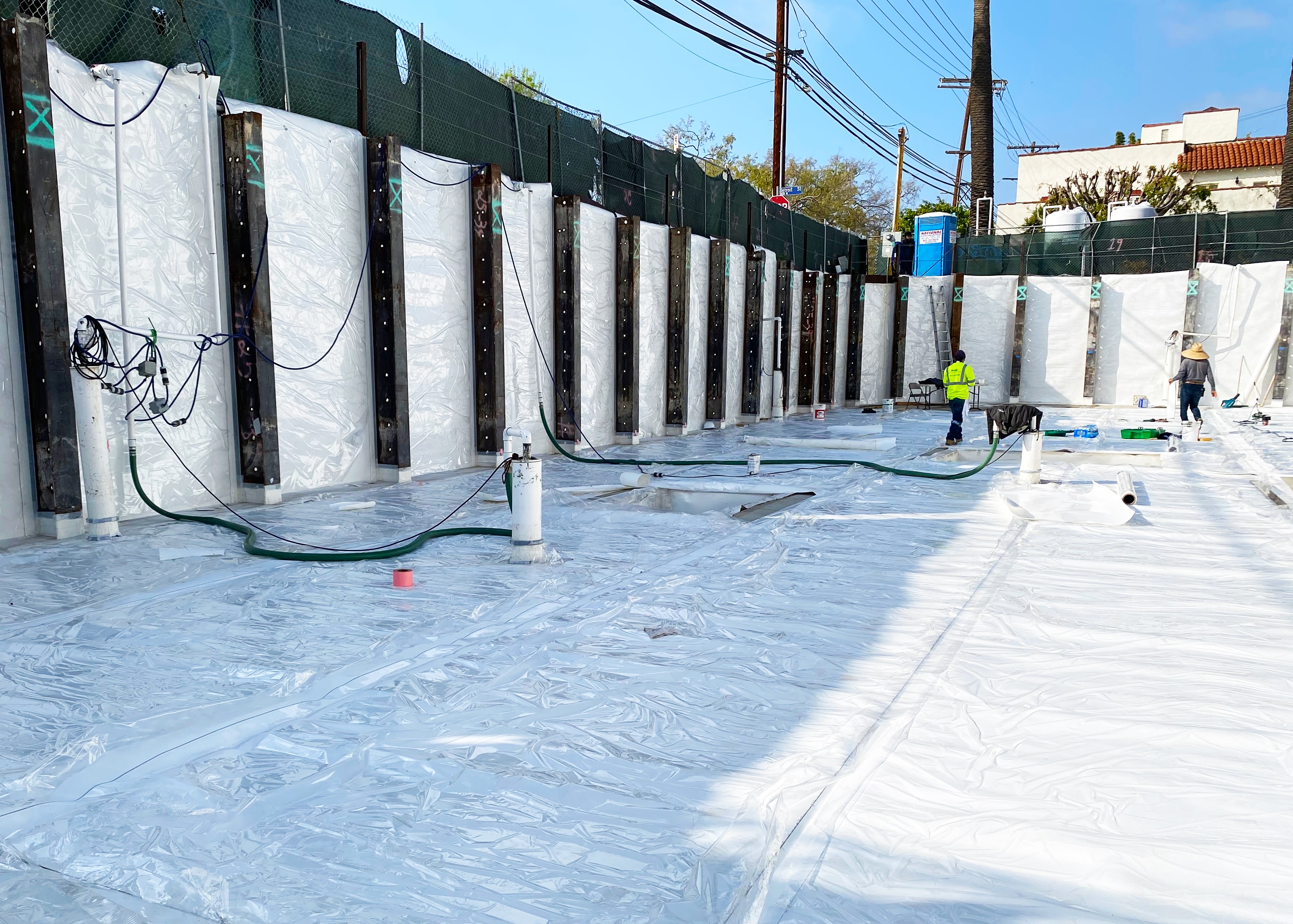 A job site has a white waterproofing material. There are two people on the job site with a blue sky and power lines also in the image.